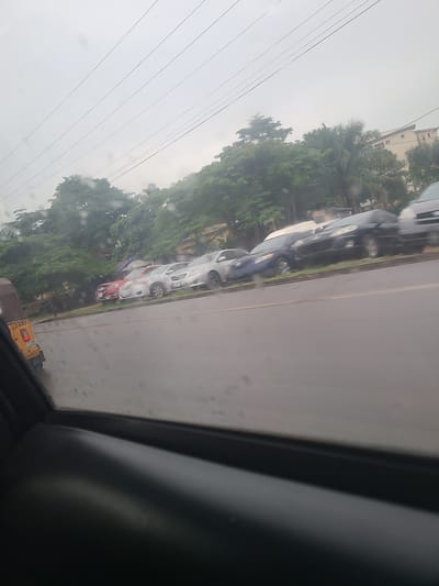 The image shows a street scene in Karu, Nigeria, viewed from a car window. Several cars are parked in a line along the side of a road. The weather appears to be overcast or rainy, as indicated by water droplets on the window. Lush green trees and some buildings are visible in the background. The overall atmosphere is one of a typical day in a busy urban or suburban area.
There are no people clearly visible in the image. The primary objects are the cars and the surrounding environment. The scene depicts a street with parked vehicles, suggestive of everyday traffic or a residential area. The presence of power lines in the sky further reinforces the urban setting.
Notable details include the overcast sky, suggesting it might be daytime, but with limited sunlight. The water on the window could indicate recent or ongoing rain. The trees suggest a tropical or subtropical climate. The presence of multiple cars lined up could be due to parking or a traffic jam, though the cars appear stationary.
A small yellow vehicle with the license plate "LZ4844" is visible towards the bottom right of the image. No other discernible text is present. The angle of the photo, being taken from inside a vehicle and tilted, gives a dynamic perspective to the scene.