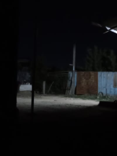 The image depicts a dark, outdoor scene, possibly a rural or industrial area in Taraz, Kazakhstan. In the foreground, a dark, vertical element, possibly a pillar or wall, is on the left side, obscuring part of the view. The ground appears to be dirt or gravel, with some lighter patches suggesting reflections or illumination. In the mid-ground, a wooden fence or wall runs horizontally across the scene. Hanging on this fence are two large, rectangular objects. The one on the left is a rusty, brown, patterned tapestry or rug. To its right is a section of light blue wooden slats. Behind the fence, dark trees or foliage are visible against a black night sky. A vertical pole, possibly a utility pole or a signpost, stands to the left of the tapestry. A ladder-like structure leans against the fence to the left of the tapestry. The overall lighting is dim, creating a nighttime atmosphere.