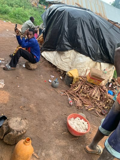 In Buga da Gindi, Nigeria, a young Black male sits on a rough-hewn log in a dusty, outdoor environment. He is wearing a blue long-sleeved shirt with red and white accents, black pants with yellow markings, and black and white patterned sandals. In his right hand, he holds a mobile phone, and his left hand rests on his head. Behind him, another adult Black male, wearing a light-colored long-sleeved shirt, is partially visible, as is a small child in a pink garment.

The immediate surroundings are characterized by packed dirt, scattered debris including plastic wrappers and pieces of paper, and a pile of dry, reddish-brown plant matter. A red bowl filled with a white, granular substance sits on the ground near the pile. To the left, a large, rough-hewn tree stump is present, with an orange plastic jug resting on the ground nearby. In the background, a makeshift structure is visible, covered by a large black plastic sheet, with parts of what appears to be corrugated metal roofing showing. Several other individuals, mostly Black males, are partially visible to the right of the frame, one of whom is wearing blue shorts. The overall impression is one of a humble, perhaps temporary, settlement.