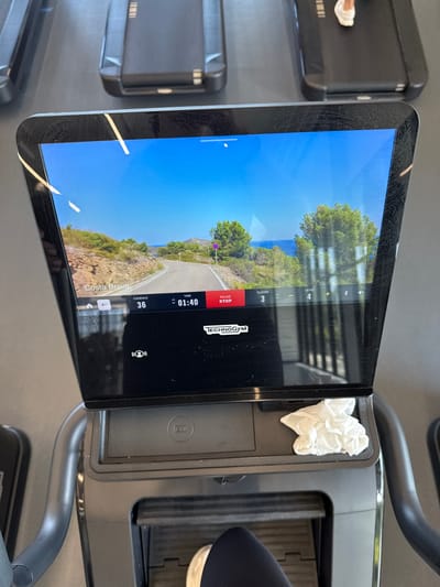 A treadmill monitor is playing a video of Costa Brava. The screen shows various statistics, including cadence (36), time (01:40), and floor (3) level (4) . The touchscreen displays the Technogym logo and has a pause/stop button. To the side, there is a white tissue. There is a person using the treadmill, with a foot visible on the right side of the image. The image was likely taken in a gym, potentially in College Station, United States of America.