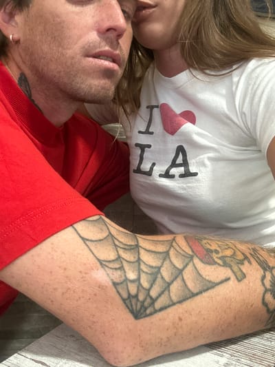 The image shows a close-up of two people. The person on the left is wearing a red shirt and has tattoos on their arm. The person on the right is wearing a white t-shirt with "I ❤️ LA" printed on it. Their faces are close together, suggesting intimacy. The background is blurred, but appears to be an indoor setting with a wooden surface. There is no indication of the location being Palma, Spain.