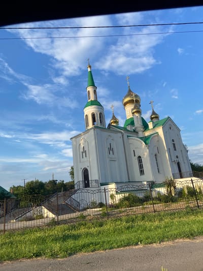 The image depicts an Orthodox church with white walls and green roofs, topped with golden domes. The church is situated in a rural or suburban setting, with a grassy area and a metal fence bordering the property. A paved road is visible to the right, and a telephone pole and wires are present in the foreground. The sky is a clear blue with scattered white clouds, suggesting a daytime scene, likely in the late afternoon or early evening due to the warm lighting. There are no people visible in the image. The overall impression is one of peacefulness and architectural beauty. The image is oriented vertically, with the church structure running from the bottom left to the top right of the frame.