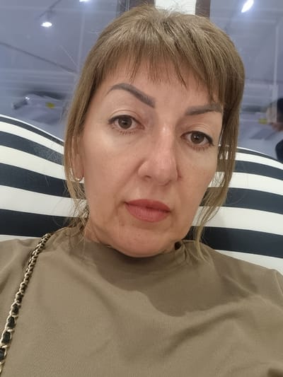 A woman with blonde hair and brown eyes, wearing a beige t-shirt and a gold chain purse, is pictured from the chest up, looking at the camera. She is sitting in front of a striped cushion. The background appears to be indoors, possibly a public space with fluorescent lighting and a large window.