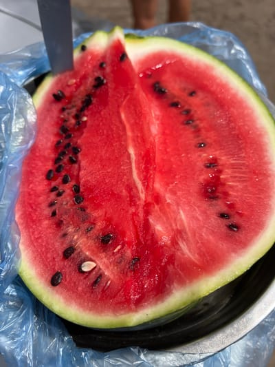 A close-up shot captures a freshly cut half of a watermelon, its vibrant red flesh speckled with black seeds, resting in a silver bowl. A portion of a knife is visible, protruding from the left side of the watermelon, indicating it has been recently cut. The watermelon is partially enclosed in a crinkled blue plastic bag, suggesting it might be prepared for transport or storage. In the background, out of focus, a person's legs are visible, implying a casual outdoor setting, possibly a picnic or a day at the beach. The ground appears sandy or dusty, and the overall lighting suggests it's daytime. The image does not provide specific details about the location within Alexandrovka, Russia.