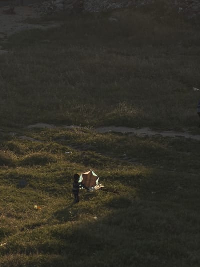 The scene appears to be an aerial view of an open, grassy area near a path. A person stands near a makeshift shelter with what looks like an umbrella-like cover. There are scattered objects nearby. The terrain appears to be uneven. The specific location within Jabalia, Palestinian Territory, is not discernible from the image.