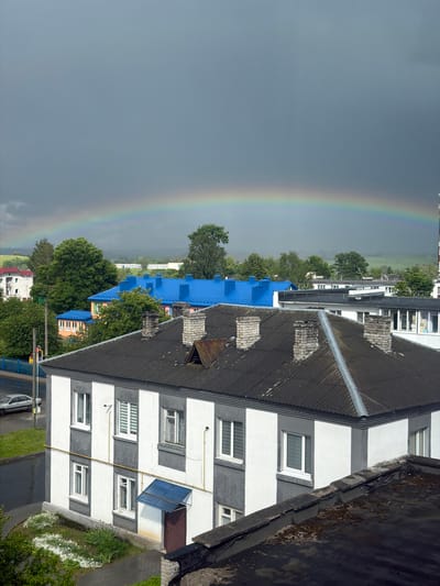 An aerial view captures a residential area in Valozhyn, Belarus, under a dramatic sky with a rainbow. In the foreground, a two-story building with a dark, corrugated roof is prominent. This building has a distinctive facade with alternating vertical bands of white and gray. It features multiple white-framed windows, some with blinds. A small blue awning shelters a brown door on the right side of the building.

Further back, a row of buildings with bright blue roofs suggests a communal or institutional setting. These buildings are interspersed with lush green trees. In the distance, more buildings and rolling, green hills are visible under a dark, overcast sky, partially obscured by mist or rain. A faint rainbow arcs across the sky, spanning from the left to the right of the frame, hinting at recent or ongoing precipitation. A silver car is parked on the wet asphalt road to the left of the foreground building. The overall scene depicts a somewhat subdued urban landscape, illuminated by the striking natural phenomenon of a rainbow.