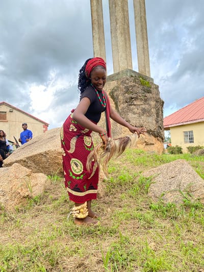 The image shows a young Black woman standing on a grassy hillside next to large rocks, possibly a monument given the concrete pillars in the background. She is wearing a black top, a red headscarf, and a vibrant red skirt with a repeating circular pattern. She has a necklace made of red and green beads. Her hair is braided and black. She is holding a strand of long, blonde and brown hair, possibly a wig or hair extension, in her right hand and smiling broadly. On her right ankle, she has a decoration made of woven yellow material. In the background, a building with a red roof and light yellow walls is visible, along with other people in casual clothing. The sky is overcast and cloudy, suggesting it might be daytime. The setting appears to be outdoors, with a mix of natural rock formations and some man-made structures. There is some text visible on a band around the woman's skirt, but it is too small to read clearly.