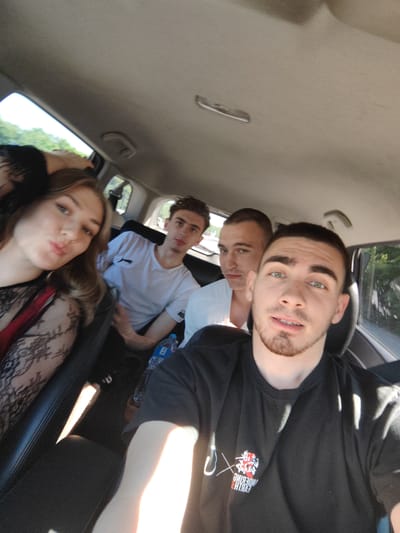 The image captures a group of four young adults inside a car, appearing to be on a road trip. The photograph is taken from the perspective of someone in the front passenger seat, looking towards the back. In the foreground, a young man with short dark hair, a beard, and green eyes is looking directly at the camera with a slight smile. He is wearing a black t-shirt with a red and white logo on the left chest. To his left, a young woman with long blonde hair is pouting her lips and looking towards the camera. She is wearing a black lace top with a red strap visible. Behind her, a young man in a white t-shirt is looking towards the front of the car, while another young man in a white t-shirt is in the middle row, facing the camera with a neutral expression. A bottle of water is visible on the seat between the young woman and the man in the white t-shirt. The interior of the car is visible, with sunlight streaming in from the windows, indicating it is daytime. The background outside the car window shows greenery, suggesting a rural or suburban setting. The image does not provide any specific visual cues to pinpoint the location in the city of Mirny, Russia.