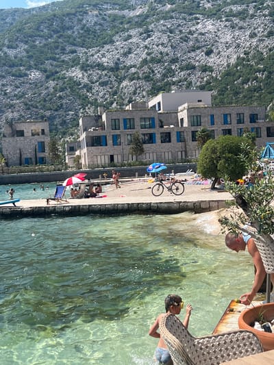 The image depicts a sunny day at a waterfront location in Risan, Montenegro. In the foreground, clear turquoise water laps against a stone quay. A young child wearing goggles and patterned swim trunks stands in the water, facing away from the viewer. To the right, a bald man in a blue tank top leans over, holding a yellow object, possibly a piece of fruit, towards the child. A woven chair is placed in the foreground, obscuring part of the scene.

In the middle ground, a paved area serves as a beach, with several people lounging on towels and sunbeds. Red and white and blue umbrellas provide shade. A bicycle is parked on the paved area. Further back, modern buildings with gray stone facades and blue-framed windows are visible, nestled at the base of a steep, rocky mountainside covered in sparse vegetation. The mountainside rises dramatically behind the buildings. Several people are seen swimming in the water near the quay. The overall atmosphere is one of a relaxed summer vacation.