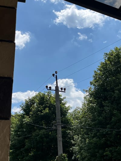 The image shows a concrete utility pole with electrical insulators and wires against a bright blue sky with scattered white clouds. Lush green trees are visible on either side of the pole, and the top right corner of the frame is partially obscured by a dark, angled overhang, possibly part of a building. The left side of the image features a portion of a building's facade, constructed with alternating bands of light brown and dark brown or black material, suggesting a textured or tiled surface. The lighting indicates it is daytime, likely midday, given the bright sun and clear sky. The overall scene depicts an outdoor environment in a city, with elements of infrastructure and nature coexisting. The specific location of Koroviia, Ukraine, is mentioned in the context of this scene.