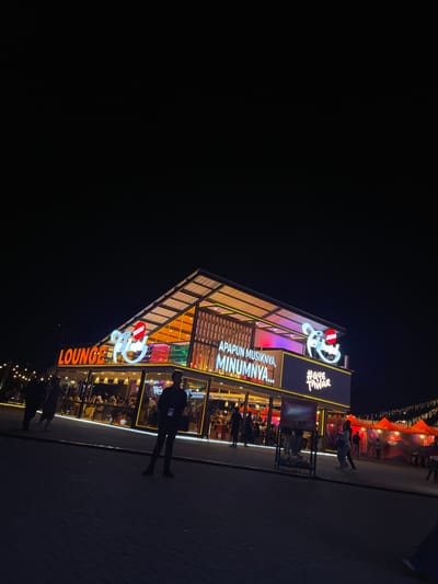 At night in Salembaran, Indonesia, a brightly lit establishment, labeled "LOUNGE" with an upper-level sign reading "APAPUN MUSIKNYA, MINUMNYA..." and "#GUETANAK", stands out against the dark sky. The building has an open, multi-level design, with patrons visible inside through glass walls. A person stands on the sidewalk outside, their shadow stretching onto the pavement. In the upper right, a partially visible, tent-like structure is illuminated. The environment is dark with a paved surface extending from the building.