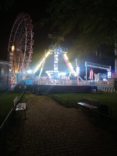 This is a nighttime photograph of an amusement park in Bessarabka, Ukraine. In the background, a large Ferris wheel with red gondolas is visible, along with several amusement rides illuminated by bright lights. The rides feature a variety of colors and structures, including a central ride with a tilting arm and a smaller rocket-shaped ride. In the foreground, a brick pathway leads through a grassy area. Two park benches are situated along the pathway, and a fence encloses the area. Trees with dark foliage are overhead, framing the scene against the night sky. The lighting suggests the park is open and operational.