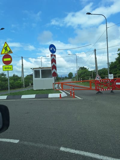 The image depicts a road leading to a checkpoint with a barrier in the village of Nigvziali, Georgia. The barrier is made of orange metal with red and white reflective markings. A small white guardhouse is situated to the left of the barrier. Several traffic signs are visible, including a "road works" sign, a "no entry" sign, and a sign indicating "600m" ahead. Two streetlights are visible in the background, along with power lines and a blue sky with scattered clouds. The road surface is asphalt, and there are white lines marking the lanes. The overall scene suggests a controlled access point on a rural road.