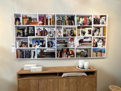 This media file presents an interior shot of a thoughtfully arranged display, potentially located in Noordwijk, Netherlands. The central subject is a large, white rectangular shelving unit mounted on a light-colored wall. This unit is segmented into numerous compartments, each showcasing an artful collection of books and magazines.
The objects on the shelves are predominantly publications, with many covers featuring iconic figures from art, fashion, and music. Visible text identifies magazines like "Vogue," "Esquire," and "New York," alongside books dedicated to artists and photographers such as Salvador Dalí, Jean-Michel Basquiat, Picasso, David Bailey, and Helmut Newton. Music-related items reference bands like The Who, U2, The Beatles, and artists like Bob Dylan and Madonna. Several covers depict David Bowie. A small, lit candle in a glass container is nestled among some books.
Below the shelving unit, a light-toned wooden cabinet or credenza is positioned. It has two closed sections and an open central compartment containing a light grey cushion. On the cabinet's surface, two white, textured rectangular boxes are placed to the left, while two white cylindrical candle holders sit on an irregular dark-colored tray to the right. No activity is taking place; the scene is a static presentation. The overall aesthetic suggests a curated space with an emphasis on visual arts, photography, and popular culture.