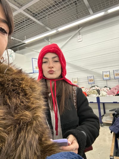 A person with brown hair wearing a red hooded sweatshirt and a black jacket poses with pouting lips. The individual is indoors, possibly in a store with price tags visible in the background. The image is angled, showing a ceiling with fluorescent lighting and a second person partially visible in the lower portion of the frame, wearing a furry coat. A hand holding a phone is also visible, with a ring on one finger.