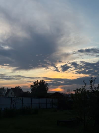 The image depicts a rural landscape at dusk in Emel'yanovo, Russia. The scene is dominated by a dramatic sky filled with clouds, showcasing hues of blue, grey, and vibrant orange as the sun sets. A few wispy clouds are scattered across the sky, while thicker, darker clouds loom towards the left side of the frame.
In the foreground, the silhouettes of trees and houses are visible against the glowing horizon. A dark fence runs across the midground, separating a grassy area from what appears to be a residential yard. A small, dark-colored structure, possibly a bench or a piece of outdoor furniture, is also present in the yard. The overall mood is peaceful and serene, capturing the quiet beauty of twilight in a rural setting. The lighting suggests it is late evening, and the sky indicates fair weather. There is no visible text in the image.