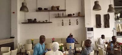The image displays the interior of a dining or lounge area located in Yola, Nigeria. The walls are predominantly off-white. The upper section of the central wall features three horizontal wooden shelves suspended by ropes, adorned with various traditional artifacts including dark bowls, terracotta pots, and carved wooden objects. Below these shelves, three metal oil lanterns are mounted on the wall. Three bell-shaped, dark pendant lamps hang from the ceiling in the foreground, with three similar lamps further back on the right. On the right wall, two large, dark wooden wall hangings, shaped like stylized spoons or paddles, are prominently displayed. A white vertical air conditioning unit is positioned on the lower right wall, and a red fire alarm pull station is visible high on the far left wall.

Approximately seven individuals are present, seated at dark wooden tables with light-colored upholstered chairs and bench seating. In the foreground left, a man with a shaved head, wearing a light blue traditional kaftan, is seated. Opposite him, the back of a person in a white hijab is visible. In the foreground right, a man wearing glasses and a patterned long-sleeved shirt is seated, smiling, with a bottled water in front of him. To his left, a young boy or short adult in a grey long-sleeved shirt is seated, facing left. In the mid-ground right, a man in a white traditional garment is seated, holding a glass. Further back, two men are seated on a beige upholstered bench; one, wearing a blue shirt, faces away from the camera, raising his right hand to form a peace sign. Tables are set with plates, food, and bottled water, indicating dining activity.