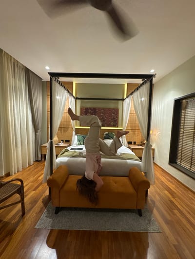 Within a well-appointed bedroom in George Town, Malaysia, a person is depicted performing an inverted pose on an orange upholstered bench. The individual, wearing a light t-shirt and light long pants, has their head positioned on the bench, with their long brown hair cascading downwards, while their legs are bent and elevated towards the ceiling. The bench, which features dark legs and tufted upholstery, rests on a light grey rectangular rug.
Behind the person and bench is a large four-poster bed with a dark metal frame draped with white sheer canopies. The bed is made with white bedding, a green bed runner, and two pillows adorned with green leaf patterns. A dark-colored laptop or tablet rests on the bed. Flanking the bed are two light-colored wooden bedside tables, each holding small objects. The wall behind the bed features horizontal light-colored woven paneling, illuminated by vertical warm-toned light strips. A rectangular artwork with a dark red and multi-colored patterned design hangs above the bed. The walls are painted a light green hue.
To the left, floor-to-ceiling sheer and cream-colored curtains cover a window. A woven chair is partially visible in the bottom left corner. To the right, a tall, narrow window with dark framing is fitted with wooden blinds. The room features polished, warm-toned hardwood flooring and a plain white ceiling with recessed lighting. A dark, blurred ceiling fan is visible in motion at the top of the frame, indicating movement.