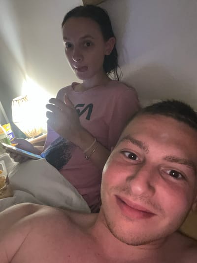 A selfie photograph shows two individuals indoors. A light-skinned male with short dark hair fills the right side of the frame, appearing closer to the camera. He has visible stubble and brown eyes, and appears to be shirtless. To the left and slightly behind him is a light-skinned female with long dark hair. She wears a pink t-shirt with a black graphic. She is holding a mobile phone. A bedside lamp illuminates the scene. In front of them, at the bottom of the frame, is food packaging. The location is unknown.