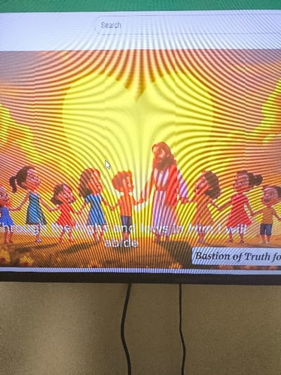 The photograph was captured in Nicosia, Northern Cyprus / Cyprus. It depicts a digital screen, likely a television or monitor, displaying an animated scene. The screen shows a central bearded figure, resembling Jesus Christ, wearing a striped robe, holding hands with approximately eight diverse cartoon-style children arranged in a semi-circle. They are positioned on a light brown surface against a radiant yellow and orange background with emanating light rays. Visible text overlays include "through the highs and lows in Him will abide" at the bottom center, and "Bastion of Truth for" within a rectangular box at the bottom right. A white "Search" bar is present at the top of the screen, and a white arrow mouse cursor points towards the center-left. Moiré patterns are visible across the screen surface. Below the screen, a textured, light beige wall is visible, with two black cables hanging vertically downwards.