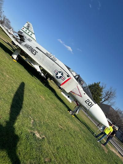 A McDonnell Douglas F-4 Phantom II fighter jet sits on green grass under a blue sky. The plane, marked "Marines" and "VMFA-333," features the number "201" on its nose and a US flag emblem. Two people stand near the jet's front end, one wearing a bright yellow shirt and the other wearing jeans. The jet is angled, and the ground slopes upwards from left to right. A building and trees are visible in the background.