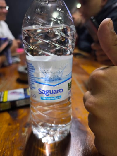 This indoor photograph, taken in Kraishte, Bulgaria, features a plastic water bottle as its central subject, resting on a textured wooden table. The image is captured at eye-level, presenting a close-up view of the bottle while the background remains softly blurred.
The primary subject is a partially consumed bottle of "Saguaro" brand table water, approximately three-quarters full. The clear plastic bottle has a distinctive spiraled, ribbed texture that reflects the ambient light. Its label is predominantly white and light blue, with "Saguaro" displayed prominently in large blue letters, and "ТРАПЕЗНА ВОДА" (Table Water in Bulgarian) printed beneath it. Smaller Bulgarian text on the label includes phrases such as "На водата" (On the water), "с обогатен" (with enriched), "минерализация" (mineralization), "от Балдаран" (from Baldaran), and expiration information.
To the right of the water bottle, a person's right hand is visible, making a "thumbs up" gesture, implying approval or positive sentiment. The hand is cropped, showing the thumb and part of the palm. To the left of the bottle, a pack of cigarettes with blue and yellow packaging lies on the wooden table.
The background, out of focus, reveals several individuals, suggesting a social gathering. One person on the far left wears glasses and a white shirt, while another in the mid-background is dressed in dark clothing. The lighting in the scene is artificial and subdued, indicating an evening or nighttime setting. The warm, rich tones of the wooden table provide a contrast to the cooler blue and white of the water bottle label. The overall impression is that of a casual and relaxed gathering in an indoor setting, likely a cafe or similar venue.