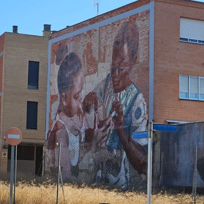 The image shows a mural painted on the brick wall of a building in Zaragoza, Spain. The mural depicts two individuals, appearing to be an adult and a child, with their hands close together, possibly engaged in an activity or interaction. The artwork has a muted color palette, with predominantly earthy tones and hints of blue and green. The building itself is made of brick, with several windows visible. To the left of the mural is another building constructed with lighter colored bricks, featuring dark-shuttered windows. In the foreground, there is a chain-link fence and some dry vegetation. A circular traffic sign with a horizontal bar is visible on a pole to the left. Street signs are also present, one reading "C Lagos de Millares" and another "C San Vicente Ferrer". The sky is clear and blue.