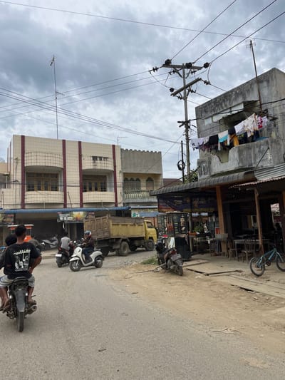 This is a street scene in Rantau Pauh, Indonesia, under a cloudy sky. A paved road occupies the foreground, with the back of a motorcycle carrying two passengers visible on the left. To the right, a white scooter with a rider is moving. Further down the road, a yellow dump truck is parked. Buildings line the right side of the street, with awnings extending over outdoor seating areas and shops. Laundry is hung on a line above one of the buildings. The architecture suggests a commercial or mixed-use area. Utility poles and numerous overhead wires crisscross the sky.
