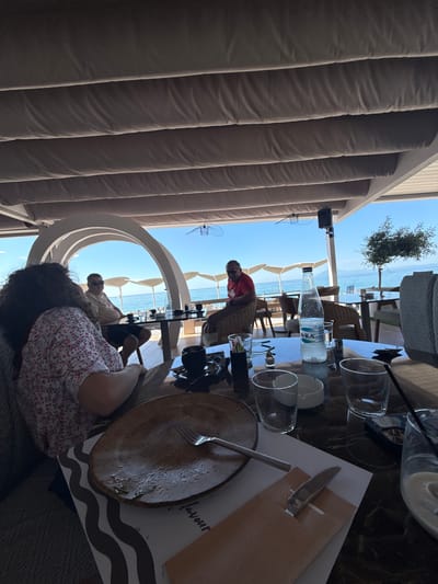 A group of four people are seated at tables under an outdoor awning overlooking the sea. The scene appears to be a restaurant or cafe in Limenaria, Greece, given the ambiance and proximity to the water.
In the foreground, a woman with long, curly brown hair is seated at a table, facing away from the camera and towards the sea. She is wearing a red and white patterned top. In front of her is a partially eaten meal on a plate, with a fork resting on it. A white napkin with a dark wavy pattern is beneath the plate.
At another table behind her, a man wearing sunglasses and a light-colored shirt is seated. He appears to be talking to someone off-camera. Further back, a man in a red t-shirt is seated at a table, leaning back in his chair. Another person is partially visible in the background.
On the tables, there are glasses, bottles of water, an ashtray, and other dining accoutrements. The awning above is made of beige fabric stretched over metal supports. Beyond the awning, the bright blue sky and the calm, turquoise sea are visible, with beach umbrellas dotting the shoreline in the distance. A small tree with green foliage stands near the water's edge. The overall impression is one of a relaxed outdoor dining experience by the sea.