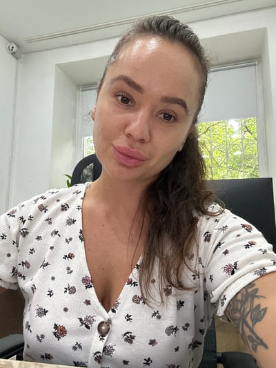 This is a portrait-oriented, low-angle selfie of a woman with brown hair and brown eyes, taken indoors. She is wearing a white, textured top with a floral print of small pink, purple, and yellow flowers and dark green leaves. The top has a V-neckline with a single brown button visible near the shoulder. She has a tattoo on her upper arm, appearing to be dark ink depicting branches and leaves, partially covered by her hair and the top.

The background includes a window with white blinds pulled partially down, revealing green foliage outside, suggesting it is daytime with trees and possibly a sunny or overcast day. To the right of the window, a dark computer monitor is partially visible. The room appears to be an office or a room with a desk, indicated by a black office chair partially in view.

The woman's face is angled towards the camera, and she is looking directly at it with a slight smile. Her makeup appears natural. A small, white, novelty object with a smiley face is visible on a white ledge in the lower left corner of the image. The lighting suggests the room is well-lit by natural light from the window. The overall mood is casual and relaxed. There is no visible text in the image.