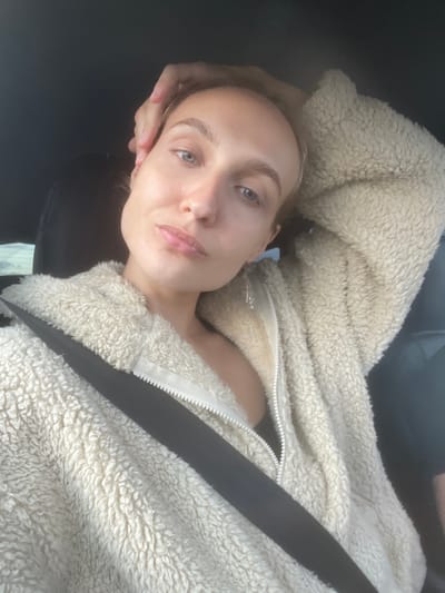 A woman is in the driver's seat of a car, wearing a fluffy cream-colored jacket with a zipper. A black seatbelt is across her chest. Her right hand is on the back of her head, and her left arm is bent, resting on her lap. The background is dark, suggesting the interior of the car. The image appears to be a selfie. The location is Pirogovo, Russia.
