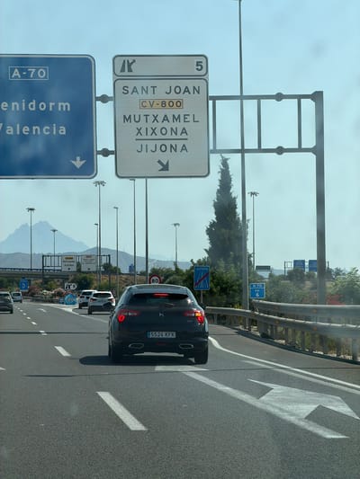 The image shows a view from a car on a highway, likely in the province of Alicante, Spain, given the road signs. In the foreground, a black DS car with license plate "5524 KFK" is being driven on a multi-lane asphalt road. White lane markings are visible, including directional arrows. To the left, another car and a blue sign with "A-70" are partially visible. In the background, other cars are seen on the road. Overhead, there are several large highway signs. One sign indicates "A-70" with destinations "Benidorm" and "Valencia" and a downward arrow. Another sign, a rectangular white panel with a grey border, lists destinations including "SANT JOAN", "CV-800", "MUTXAMEL", "XIXONA", and "JIJONA", with corresponding arrows. Further in the background, mountains are visible, along with utility poles and trees. The sky is bright and hazy.