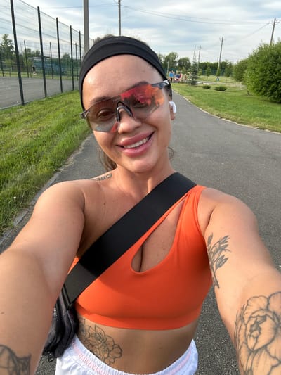 A woman with a black headband and pink-tinted sunglasses is taking a selfie while outdoors. She is wearing an orange sports bra, white shorts with pink stripes, and a black crossbody bag. There are tattoos visible on her shoulders, arms, and torso. She has white wireless earbuds in her ears and is smiling. The background shows a paved path, green grass, trees, and a chain-link fence enclosing a sports court. In the distance, there appears to be a playground and utility poles. The overall setting suggests an outdoor recreational area. The location is identified as Yuzhnouralsk, Russia.