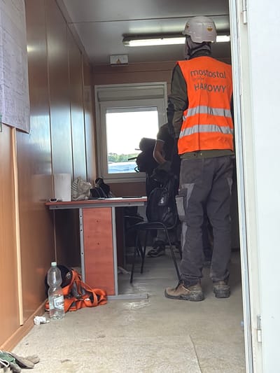 The image depicts the interior of a portable building, likely a temporary site office or changing room, at a construction location in Stróża, Poland. The scene is occupied by two individuals, both wearing work attire.
In the foreground, a person is viewed from the back, wearing a white hard hat, an orange high-visibility vest with reflective stripes, and dark work trousers. The vest has text printed on the back, including "mostostal kraków" and "HAKOWY". This person is also wearing sturdy brown work boots.
Behind this individual, another person is partially visible, also in work clothes. They are standing near a window and a desk.
The portable building has wooden paneling on the walls and a simple metal-framed desk with an orange front panel. On the desk, there are various items, including a white paper roll, a crumpled plastic bag, and what appears to be a small electronic device. On the floor next to the desk is a large plastic bottle of water and an orange safety harness. A second backpack is visible near the person in the background.
The overall environment appears functional and utilitarian, consistent with a construction site. The lighting suggests it is daytime, with natural light entering through the window and artificial light from a fixture visible on the ceiling. The floor is concrete.