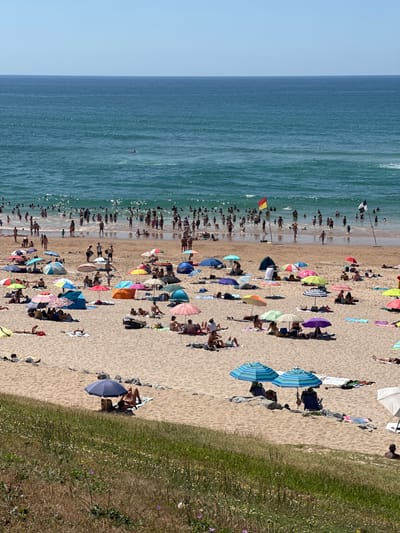 This is a photograph of a crowded beach in Biarritz, France, on a sunny day. The ocean is a vibrant blue, with small waves. Many people are in the water, swimming and playing. On the sandy beach, numerous umbrellas provide shade for sunbathers. People are scattered across the sand, some sitting, some lying down, and some walking. Beach tents are also visible. In the background, a red and yellow flag and a black and white flag are planted in the sand, likely indicating lifeguard stations or designated swimming areas. The overall scene depicts a lively and popular beach resort.