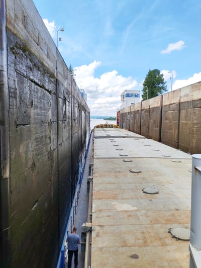 The image depicts a view from a large cargo barge moving through a canal lock. The barge occupies the right side of the frame, its deck covered in circular hatches. To the left, a massive concrete wall of the lock rises vertically, showing signs of age and moisture. In the distance, at the end of the lock, another wall is visible, with a building and a tall tree situated behind it. The sky above is partly cloudy with a bright blue hue. A single person, a man wearing a blue t-shirt and dark pants, is walking along the side of the barge, facing away from the camera. The overall scene suggests a functional, industrial environment related to water transport. The location is identified as being in Белоусово, Russia.