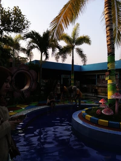 The image depicts an outdoor scene at what appears to be a water park or recreational area in Kebonsari, Indonesia. In the foreground, a young woman wearing a hijab and a beige sweater is holding a mobile phone, looking to her left. Adjacent to her, a large, stylized flower sculpture with a dark interior and red petal-like structures adorned with white spots is visible.
In the middle ground, several individuals are interacting near a pool of blue water. Some are standing or crouching around the edge of the pool, while others are seated. The pool area is surrounded by a colorful, tiled border. Palm trees are prominent throughout the scene, with their fronds extending towards the sky. One palm tree trunk on the right is wrapped with green and yellow bands. In the background, a blue building with dark windows and signage is present. Several decorative mushroom-shaped objects, red with white spots, are placed around a small island of grass. The overall lighting suggests it is late afternoon or early evening.