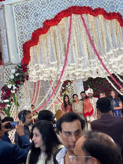 The image captures a vibrant and festive gathering, likely a wedding or a celebratory event, characterized by elaborate floral decorations and many attendees. The photo is rotated 90 degrees clockwise.

In the center of the image, serving as a grand backdrop, is a large, ornate floral arrangement. It features cascading strings of white or cream-colored small flowers, creating a textured, fan-like pattern. Interspersed within these white strands are horizontal or slightly curved lines of bright pink flowers, adding a contrasting pop of color. The outer edge of this fan-like structure is bordered by a thick, wavy garland of rich red flowers, possibly roses or carnations. Below this main floral display, there are smaller bouquets of red and white flowers with green foliage, resting on decorative white lattice-work panels. Behind the main floral backdrop, a wall is densely covered with red and green foliage, giving the impression of a lush, garden-like setting.

Numerous people are present, indicating a large social event. Towards the center-right of the image (if viewed correctly), a central couple is visible, suggesting they are the focus of the event. The man is dressed in traditional Indian wedding attire, including a white sherwani, a white turban, and a red decorative shawl or dupatta draped over his shoulder. The woman next to him appears to be wearing a light-colored, possibly golden or cream, traditional outfit. They are surrounded by other guests.

Several other individuals are seen in the foreground and background. On the far right, in the immediate foreground, a man's face is partially visible, wearing spectacles and looking towards the center. Another woman with long dark hair is also in the foreground, looking downwards. Other guests, both men and women, are dressed in formal and traditional Indian attire, such as sarees, lehengas, and formal shirts. Some attendees are holding mobile phones, seemingly capturing photos or videos of the event. One person in the lower foreground is wearing headphones and holding a camera or phone, possibly a photographer or videographer. The expressions on the faces of the visible guests range from attentive to joyful, reflecting the celebratory nature of the occasion.

The lighting appears bright and even, suggesting either a well-lit indoor venue or an event taking place during the day or early evening. The overall atmosphere is one of festivity and celebration. No discernible text is visible in the image.