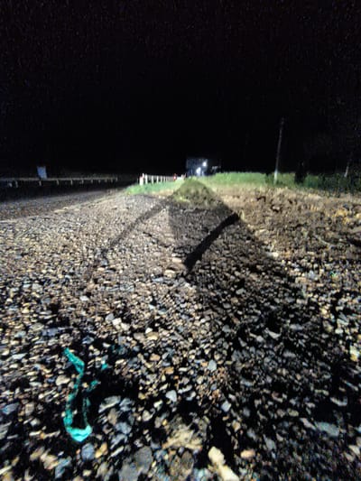 The image shows a dark, unpaved road covered in gravel and stones at night. Headlights illuminate the road, casting long shadows. In the distance, a building with lights on is visible, along with a few distant figures. There is some green vegetation on the sides of the road. In the foreground, a piece of bright green plastic debris is visible on the ground. The scene suggests a rural or undeveloped area, potentially on the outskirts of Thôn 9, Vietnam, given the context of the query. The overall impression is one of a dimly lit, possibly remote, nighttime scene.