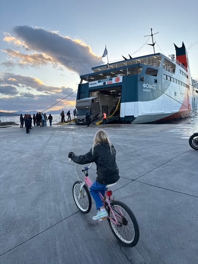 The image depicts Aegina Port, Greece, during late afternoon or early evening under a partly cloudy sky. In the foreground, a blonde-haired child, viewed from behind, rides a pink bicycle on a grey concrete pier. The child wears a dark jacket, blue pants, and light-colored sneakers, and faces a large vehicle and passenger ferry.

The ferry, identified as "PHIVOS" (ΦΟΙΒΟΣ) and "SARONIC," is docked at the pier with its stern ramp lowered, connecting to the concrete surface. The ferry's hull features dark teal, white, and red sections, and displays "ΠΕΙΡΑΙΑΣ" (Piraeus). A Greek flag flies from the ferry's mast. A grey delivery van is positioned on the ferry ramp, with an individual in an orange safety vest standing nearby. Another person in a dark uniform is also visible on the ramp. Approximately 15 to 20 individuals are gathered on the pier near the ramp, consistent with embarkation or disembarkation procedures. Distant mountains are visible across the water on the horizon. A portion of a motorcycle's front wheel is visible on the right edge of the pier.