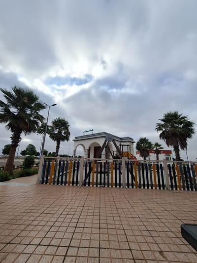 This image shows a public area with a building that appears to be a mosque, indicated by Arabic script on its facade. In front of the mosque is a children's playground with swings and a slide. Palm trees are scattered throughout the scene, and the ground is paved with square tiles. A multi-colored fence separates the playground from the paved area. The sky is overcast, suggesting a cloudy day. The location is identified as Oulad Hcine, Morocco.