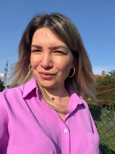 A woman with blonde hair is taking a selfie outdoors. She is wearing a pink collared shirt and large hoop earrings. The background shows a blue sky, trees, a fence, and a tall industrial tower with power lines. The woman is looking at the camera and smiling slightly. The location is identified as Yanovo-Grushevsky, Russia.