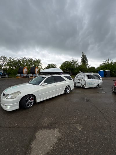 The image depicts two white vehicles parked on an asphalt surface, likely a parking lot. The sky is overcast. The vehicles are a white station wagon with a cargo carrier on top and a white truck with customized art. Utility cables on a trailer can be seen in the background along with portable toilets. The location is the city of Pavlovskaya, Russia.