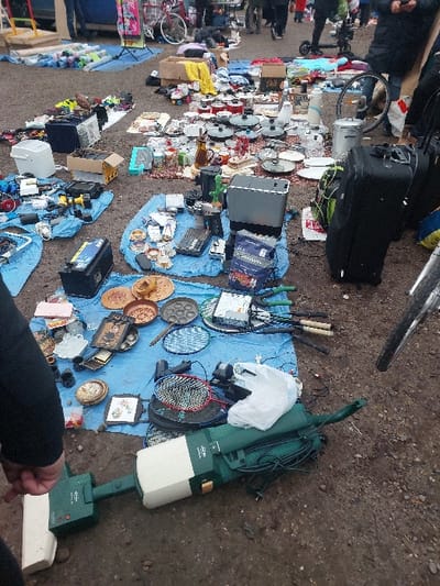 The image depicts an outdoor market scene on a dirt and gravel surface in Eckbolsheim, France. Various used items are displayed for sale on blue tarpaulins and directly on the ground. In the immediate foreground, a green and beige Vorwerk Kobold upright vacuum cleaner lies horizontally, with a white plastic bag resting on its upper section. The front wheel of a bicycle is partially visible on the far right.
The market stalls showcase a diverse array of merchandise:
- **Household Goods:** Numerous pots, pans (some with lids), ceramic plates, glassware, a decanter, an electric kettle, a coffee maker, a toaster oven, a blender, and various small decorative items are visible across several blue tarpaulins.
- **Electronics and Tools:** Items include power tools (e.g., a drill and battery charger), a car battery, a stereo receiver, and assorted cables.
- **Sporting Equipment:** Several badminton rackets are laid out on a tarp.
- **Miscellaneous:** Cardboard boxes, a white plastic bucket, and rolls of material are also present.
In the mid-ground and background, multiple individuals, primarily visible from the waist down, are seen walking through the market, suggesting active browsing. Their attire indicates cooler weather. A red bicycle and an electric scooter are identifiable among the background activity. A human left arm, clad in a dark long-sleeved garment, and a hand are partially visible in the bottom-left corner, near the vacuum cleaner. The lighting suggests an overcast sky.