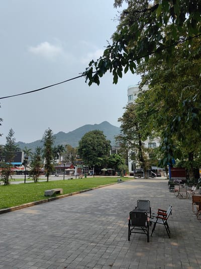 The photograph shows a paved area in Thành phố Hà Giang, Vietnam. Two sets of folding chairs are arranged on the paved surface. A strip of grass separates the paved area from a street with various commercial establishments, including a restaurant named "HAG FOODS". Beyond the street, a mountain range forms the backdrop. Trees are present throughout the scene, partially obscuring buildings and adding to the natural environment. The sky is overcast.
