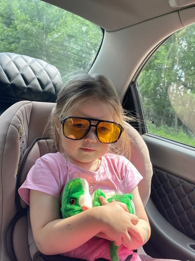 In Nekrasovo, Russia, a young girl with blonde hair is seated in a car's child safety seat, wearing large, orange-tinted sunglasses and a pink t-shirt. She is holding a green stuffed toy. The background shows trees and greenery through the car windows. The interior of the car features a black quilted panel on the door.