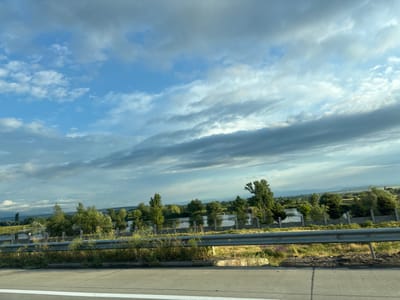 This is a landscape photograph taken from a moving vehicle on a highway, likely in the vicinity of Samtsevrisi, Georgia, given the context. The image captures a serene scene with a bright blue sky filled with scattered clouds of varying densities, some appearing dark and others white, suggesting dynamic weather patterns. In the middle ground, a body of water, possibly a lake or river, is visible, reflecting the sky and surrounded by lush green trees and vegetation. A black metal fence with decorative elements runs along the edge of the water, separating it from a grassy embankment. Beyond the water, rolling hills and more trees can be seen in the distance, with hints of mountainous terrain under the cloudy sky. In the foreground, the edge of a paved road and a reflective metal guardrail are prominent, indicating the perspective from inside a vehicle traveling on a highway. The overall impression is one of natural beauty and tranquility, characteristic of the Georgian countryside.