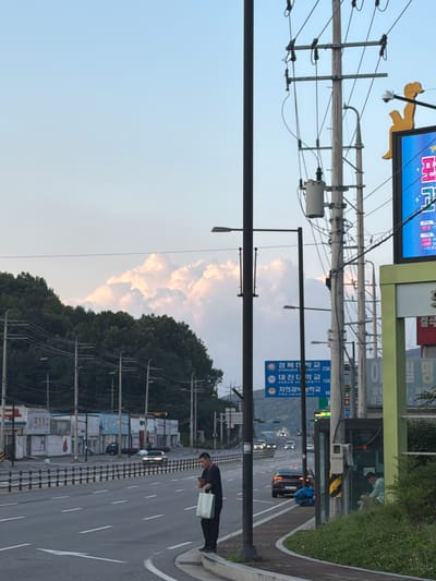 The image shows a street scene in Pocheon-si, South Korea. A man stands on the side of a road, holding a shopping bag and looking at his phone. The road is lined with buildings and trees. In the background, there are clouds in the sky. There are also several cars on the road. The image appears to have been taken in the late afternoon or early evening.