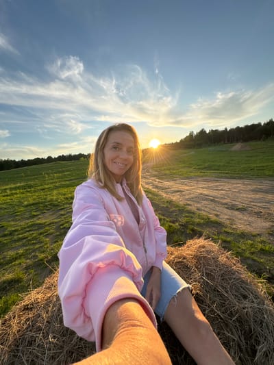The image displays a vibrant outdoor selfie, captured with a tilted perspective, showing a woman smiling brightly in a natural landscape during the golden hour. The central subject is a woman with blonde hair, wearing a light pink zippered jacket and light blue denim shorts, her bare arm visible in the foreground. She is sitting or leaning on a large, dry bale of hay or straw, which grounds the lower right portion of the frame.

The background unfolds into an expansive rural scene. Lush green fields and areas of exposed earth or dirt paths stretch towards a distant, dark line of trees, indicating a forest. The sky above is a clear, brilliant blue, adorned with delicate, wispy white clouds. The sun is a dominant feature, positioned low on the left side of the frame, emitting a powerful golden glow and a distinct lens flare, strongly suggesting either sunrise or, more likely, sunset. This warm light bathes the entire scene, highlighting the textures of the hay and the woman's attire.

The woman's cheerful smile conveys a sense of happiness and contentment, enhanced by the serene beauty of the natural setting bathed in the warm, soft light. No text is visible in the image. The scene suggests a peaceful moment in the countryside, possibly near Стеклянный, Russia, given the provided context.