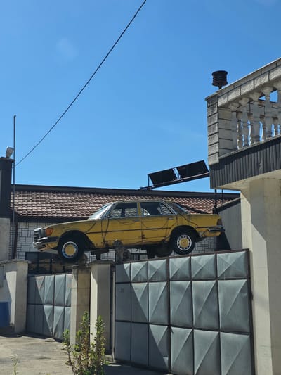 An abandoned, weathered yellow car rests precariously on top of a grey metal gate in Bregovo, Bulgaria. The car, a sedan model with visible rust and damage, is angled with its front end higher than its rear. The gate, which appears to be made of corrugated metal panels with a diamond pattern, is closed. Behind the gate and car, a tiled roof with brown tiles is visible, along with the side of a light-colored building. A white balustrade with decorative pillars runs along the top of another building to the right, and a dark, conical siren-like object sits on a pillar above the balustrade. The sky is clear and bright blue. A power line stretches diagonally across the upper portion of the image. The scene is bathed in bright sunlight, casting shadows.